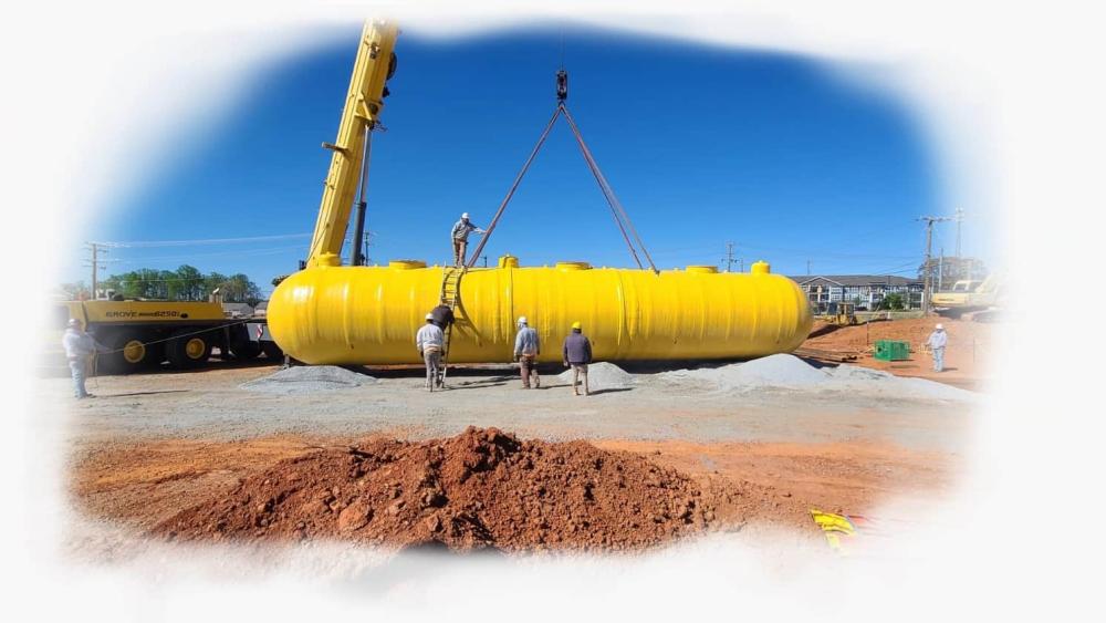 Underground fiberglass tank is being lowered into an excavated hole by a crane.
