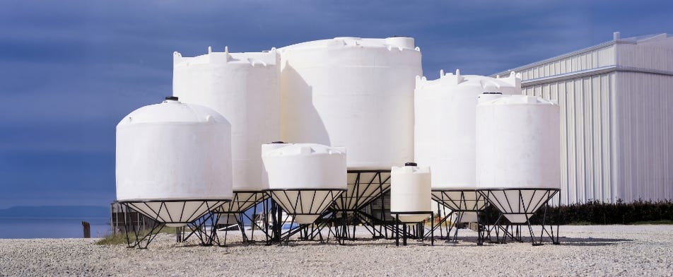 Group of white cone-bottom industrial storage tanks in multiple sizes mounted on black steel support stands outside a metal facility. Group of white cone-bottom industrial storage tanks in multiple sizes mounted on black steel support stands outside a metal facility.
